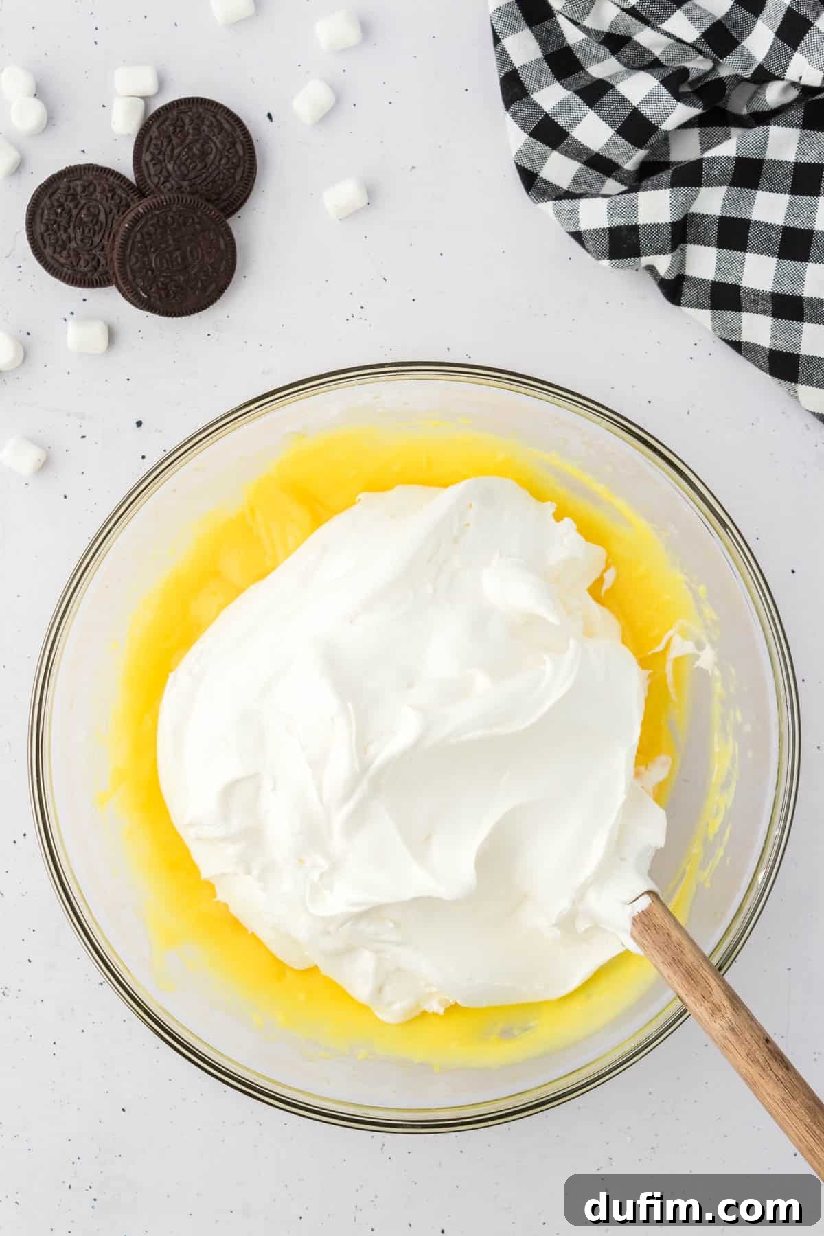 No-Fuss Oreo Fluff 6 Creamy vanilla pudding in a glass bowl, now topped with a generous dollop of cool whip, surrounded by Oreos, mini marshmallows, and a black and white checkered kitchen towel, illustrating the addition of whipped topping.