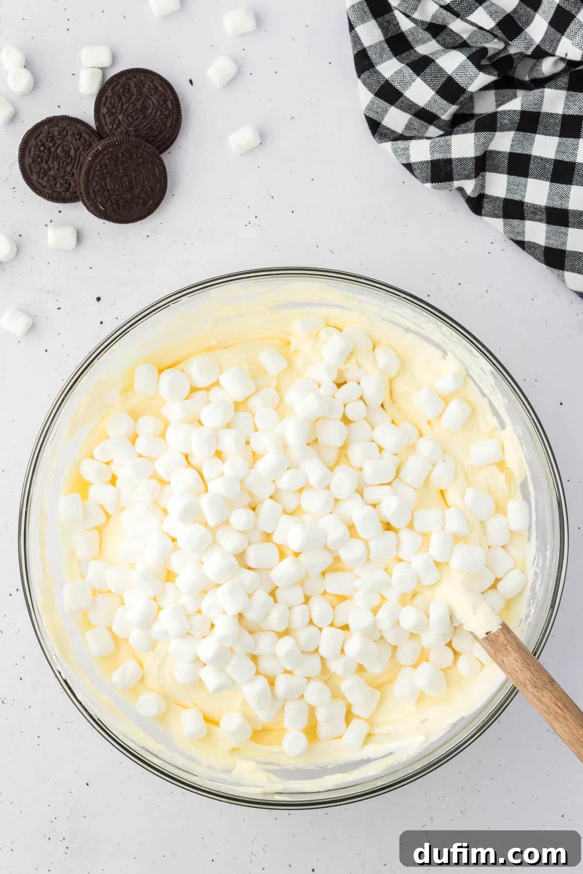 No-Fuss Oreo Fluff 7 The vanilla pudding mixture now featuring delightful mini marshmallows, contained within a glass bowl and surrounded by whole Oreos, extra mini marshmallows, and a stylish black and white checkered kitchen towel.