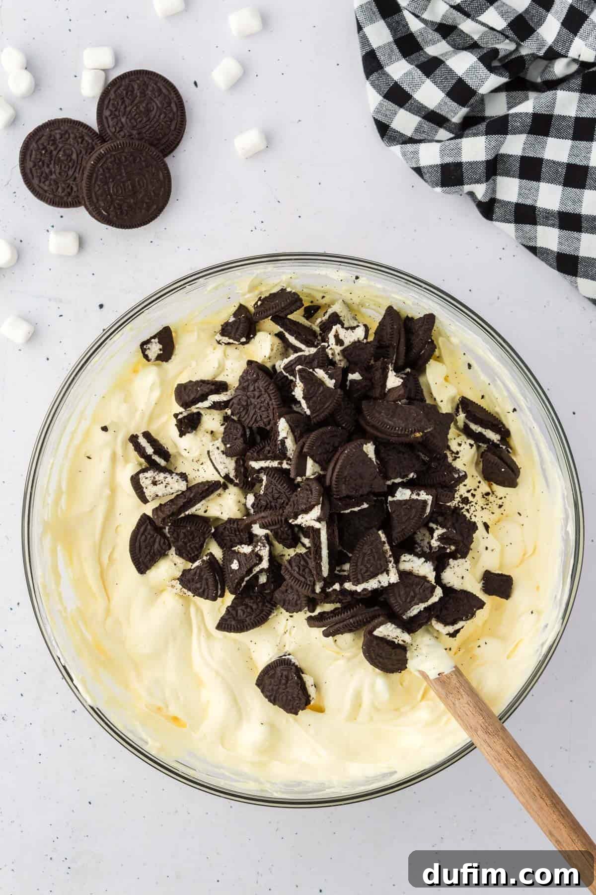 No-Fuss Oreo Fluff 8 A bowl of delicious Oreo fluff mixture, generously topped with an extra layer of chopped Oreos, and artfully surrounded by whole Oreos, mini marshmallows, and a black and white checkered kitchen towel, highlighting its readiness.