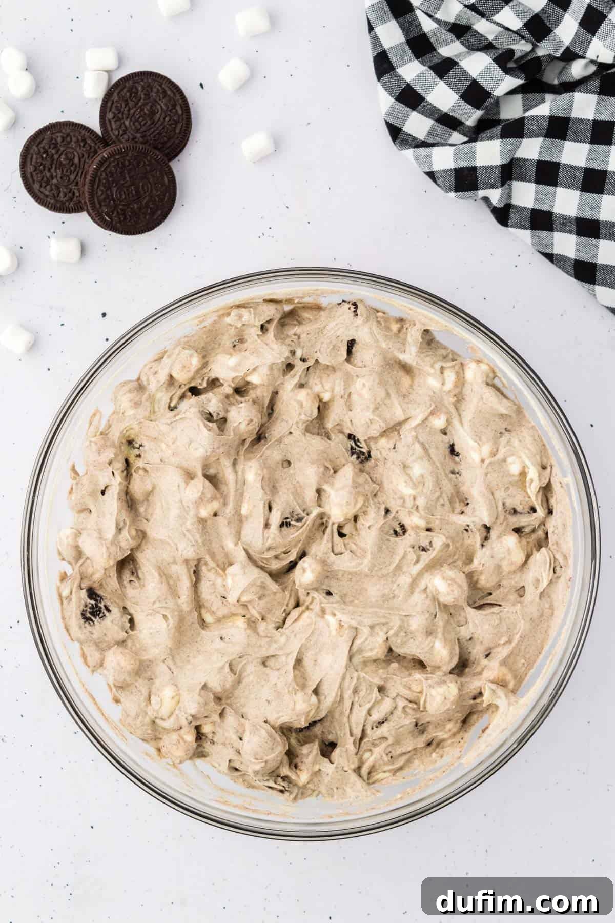 No-Fuss Oreo Fluff 9 A full glass bowl of creamy Oreo fluff, elegantly presented alongside whole Oreos, more mini marshmallows, and a black and white checkered kitchen towel, ready for serving.