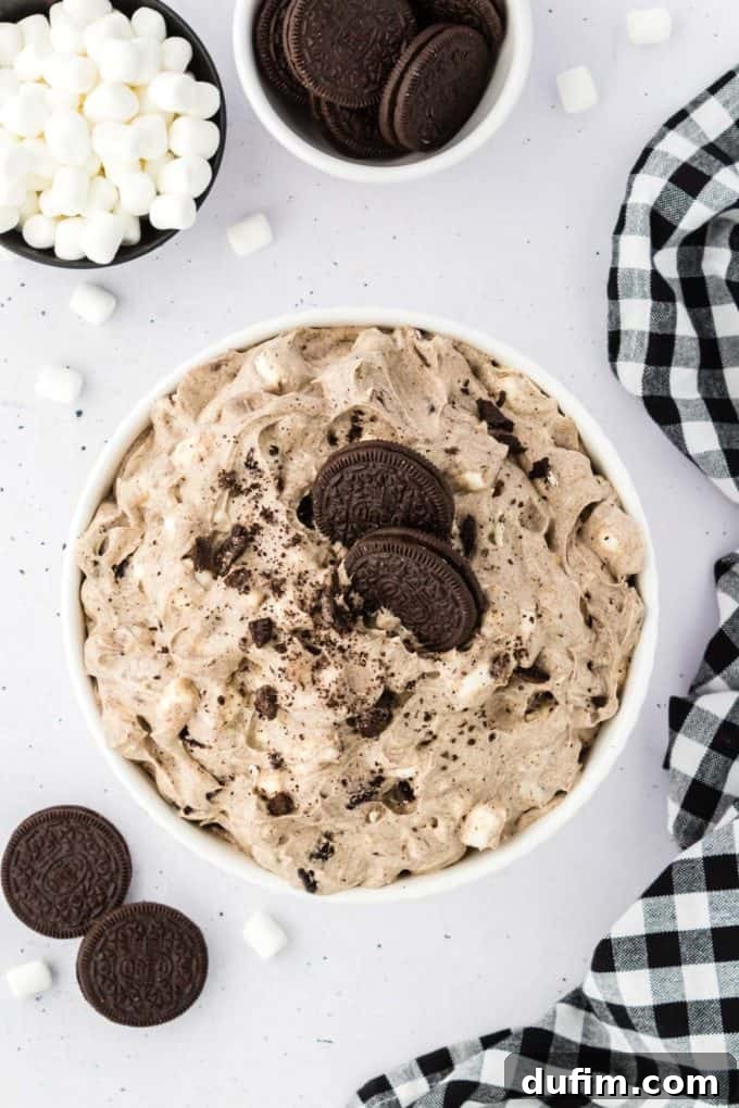 No-Fuss Oreo Fluff 10 A close-up of Oreo fluff, beautifully topped with a sprinkle of Oreo crumbs and two pristine whole Oreos, presented in a glass bowl, with additional Oreos, mini marshmallows, and a black and white checkered kitchen towel nearby.