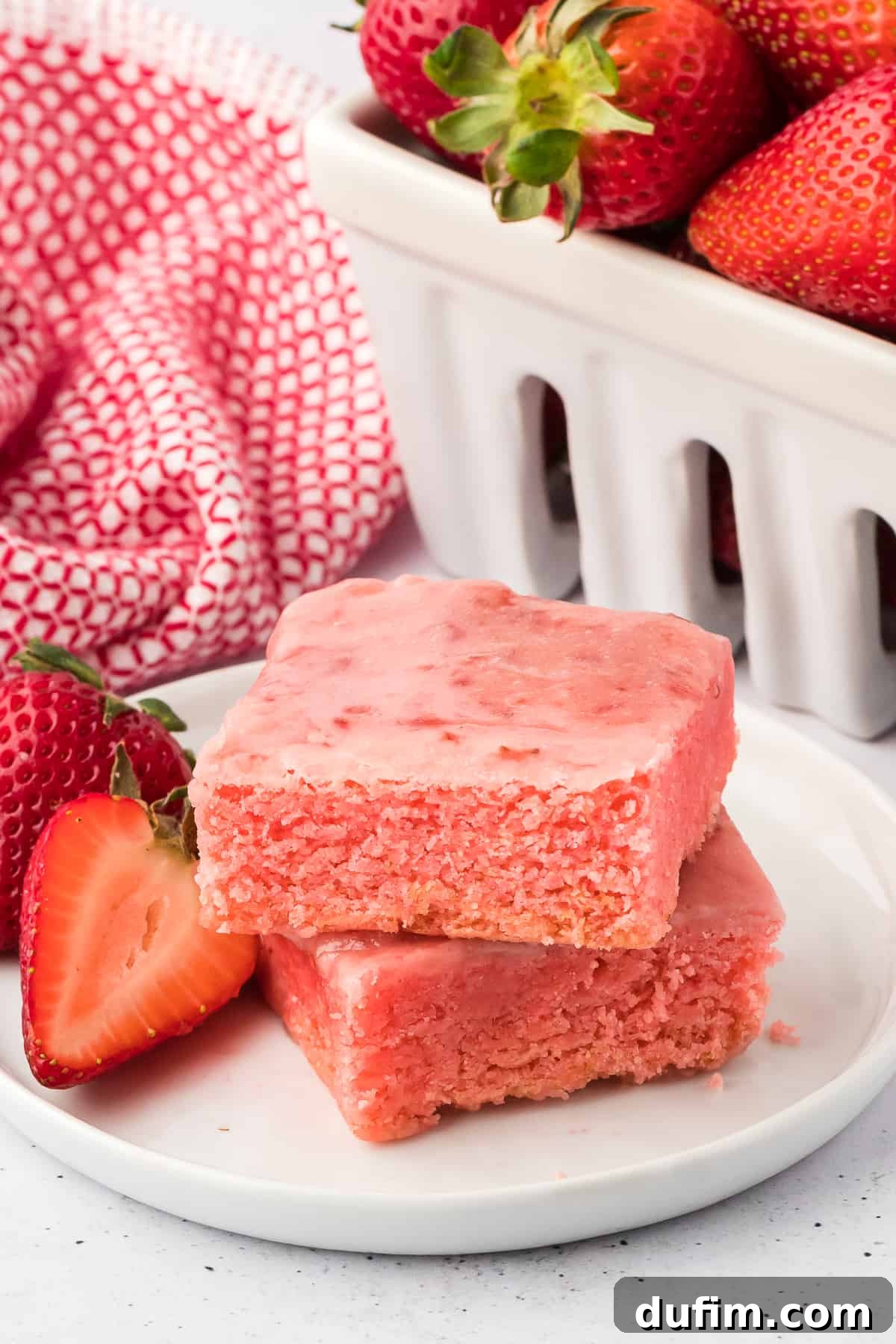 two strawberry brownies stacked on a white plate with fresh strawberries beside them surrounded by a red and white kitchen towel and a white basket of more whole fresh strawberries