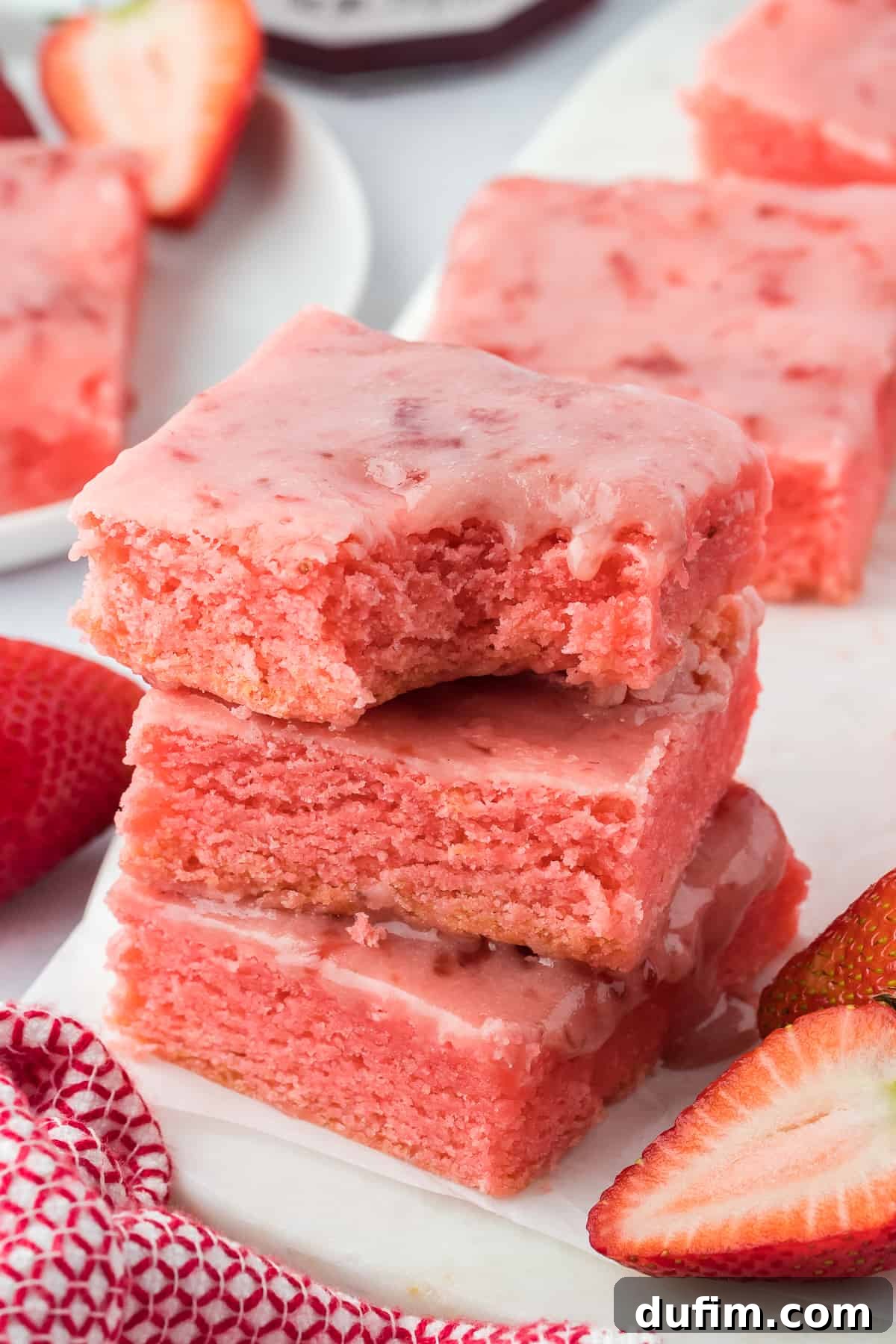 three strawberry brownies stacked with a bite out of the top brownie, a red and white kitchen towel beside them and fresh strawberries all around, and more brownies in the background