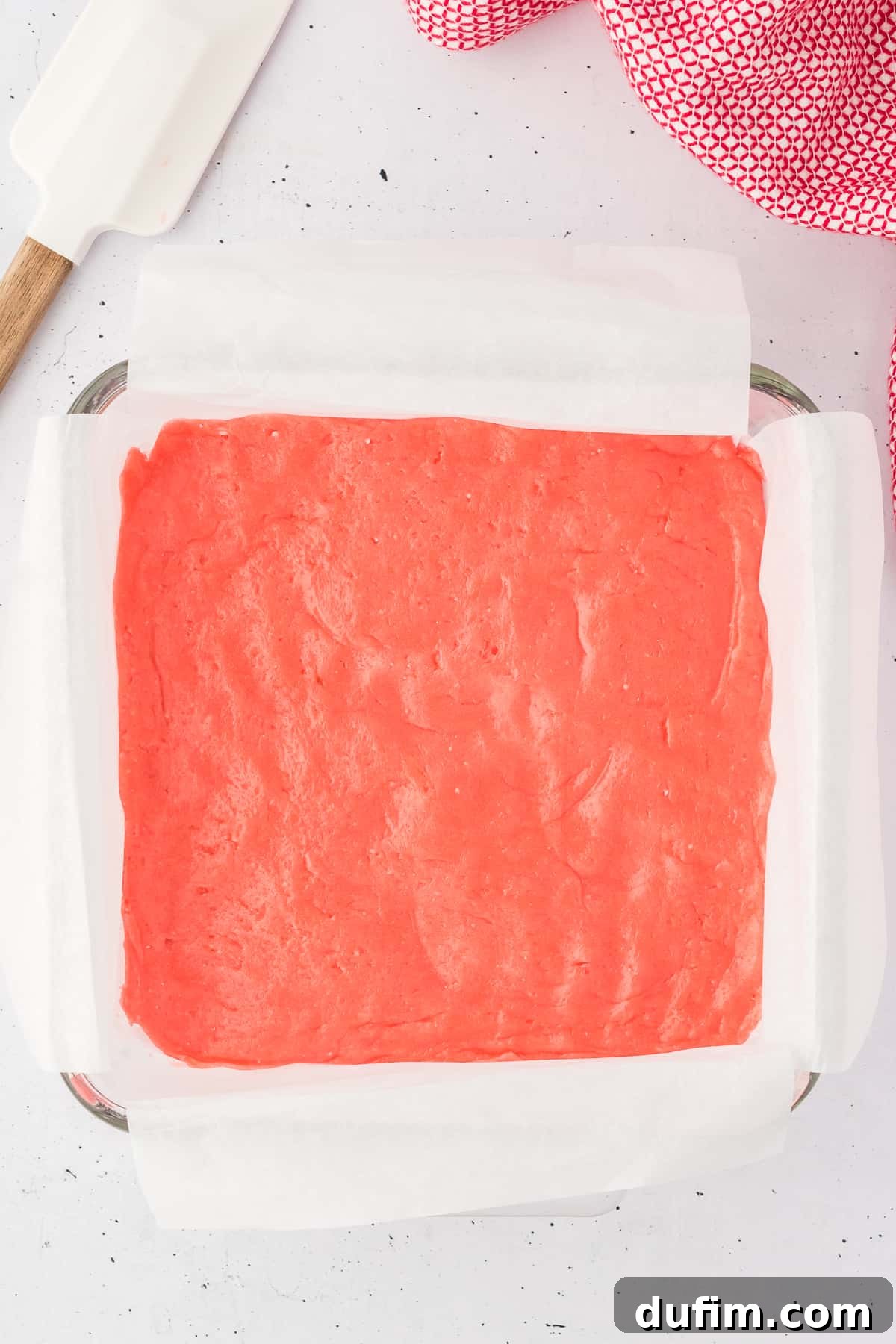 strawberry brownie batter in a glass pan lined with parchment paper surrounded by a white spatula and a red and white kitchen towel