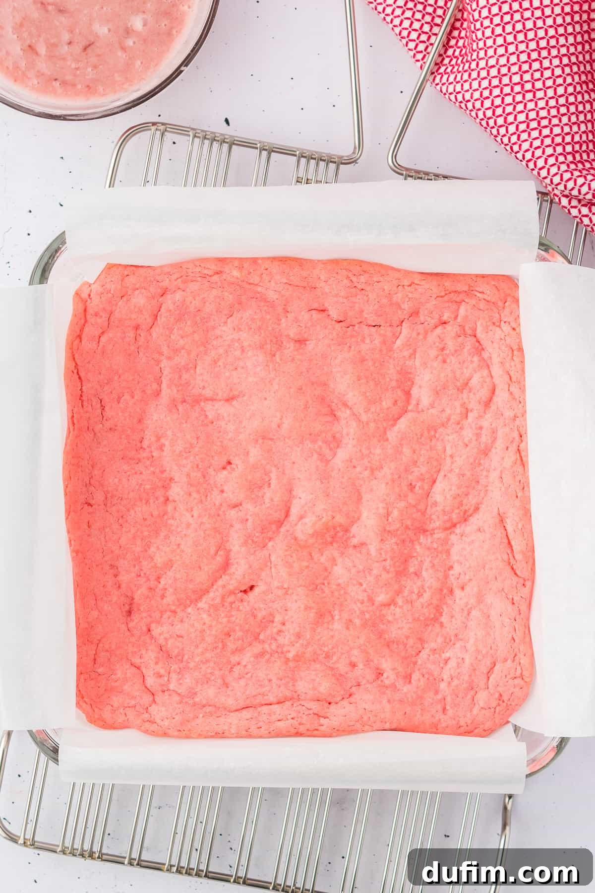 a pan of strawberry brownies on a wire cooling rack surrounded by a red and white kitchen towel and a bowl of brownie batter