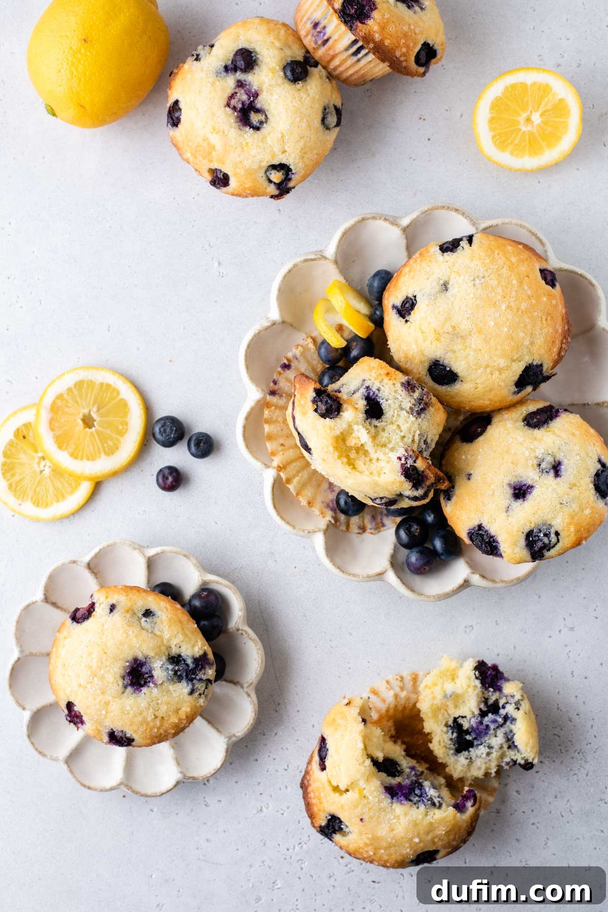 Several lemon blueberry muffins arranged on small plates and on a counter with lemon slices, lemon peels and blueberries sprinkled around them