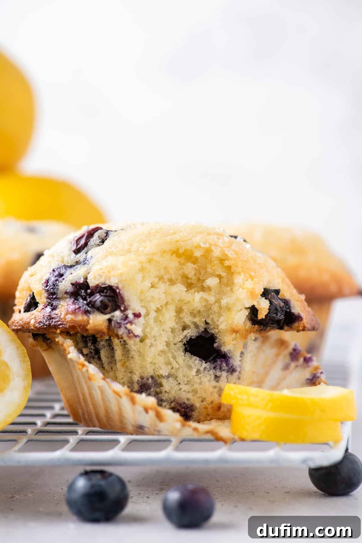 Close up of the side of a lemon blueberry muffin with one bite taken out, sitting on top of a wire cooling rack with a lemon peel beside it