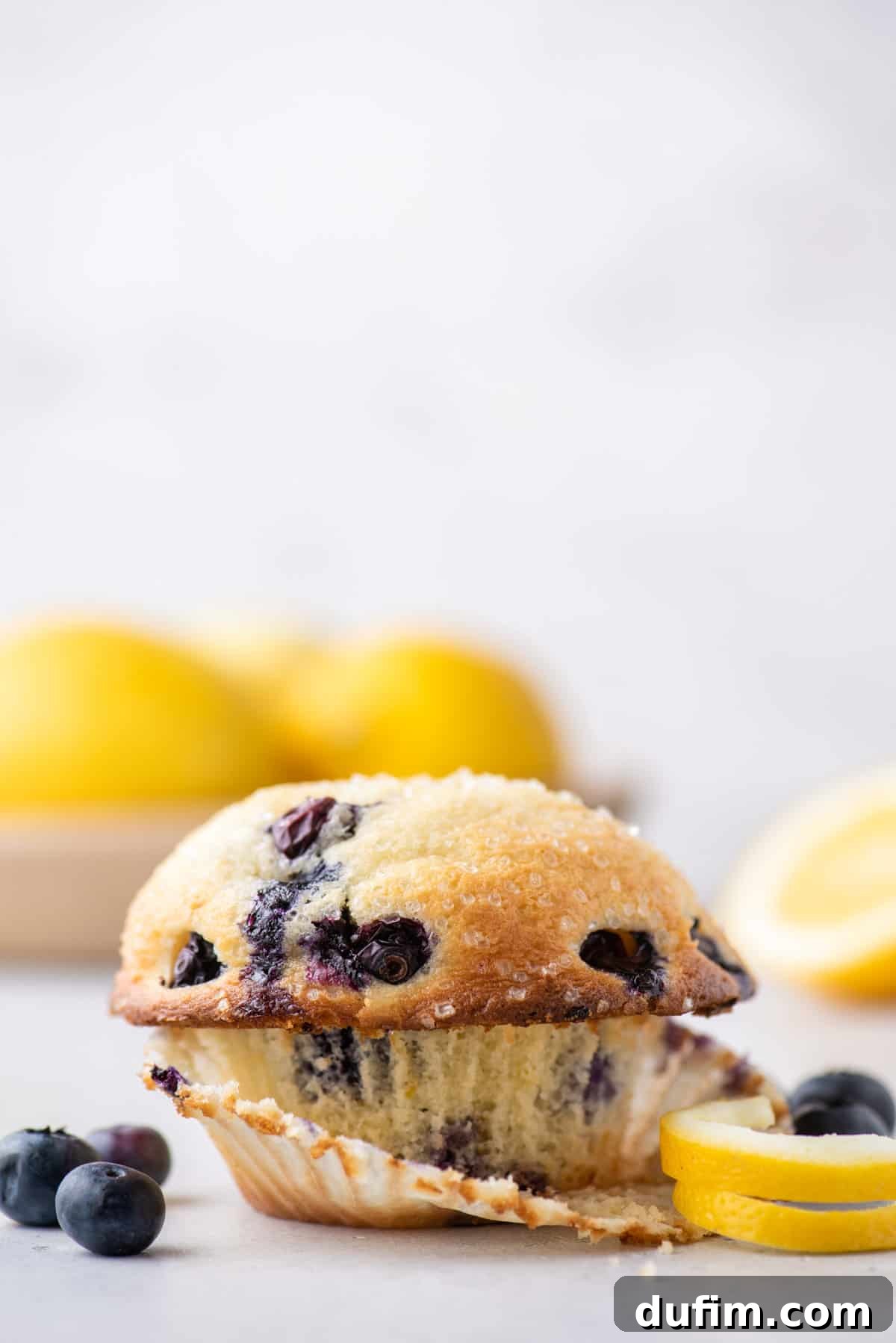 A bakery style lemon blueberry muffin with the muffin liner partially peeled away surrounded by lemon peels and fresh blueberries