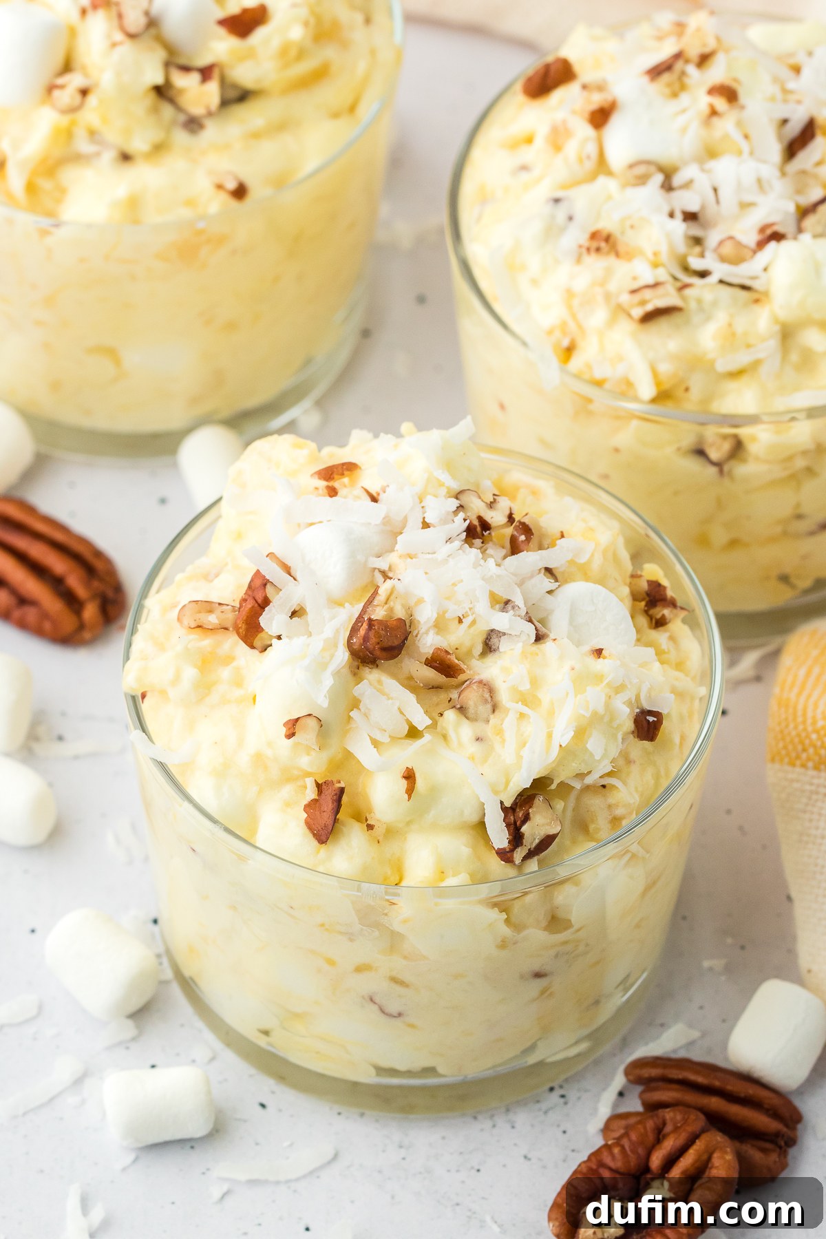 Three clear trifle dishes beautifully filled with pineapple fluff, garnished with a sprinkle of nuts, mini marshmallows, and shredded coconut, highlighting its elegant presentation.