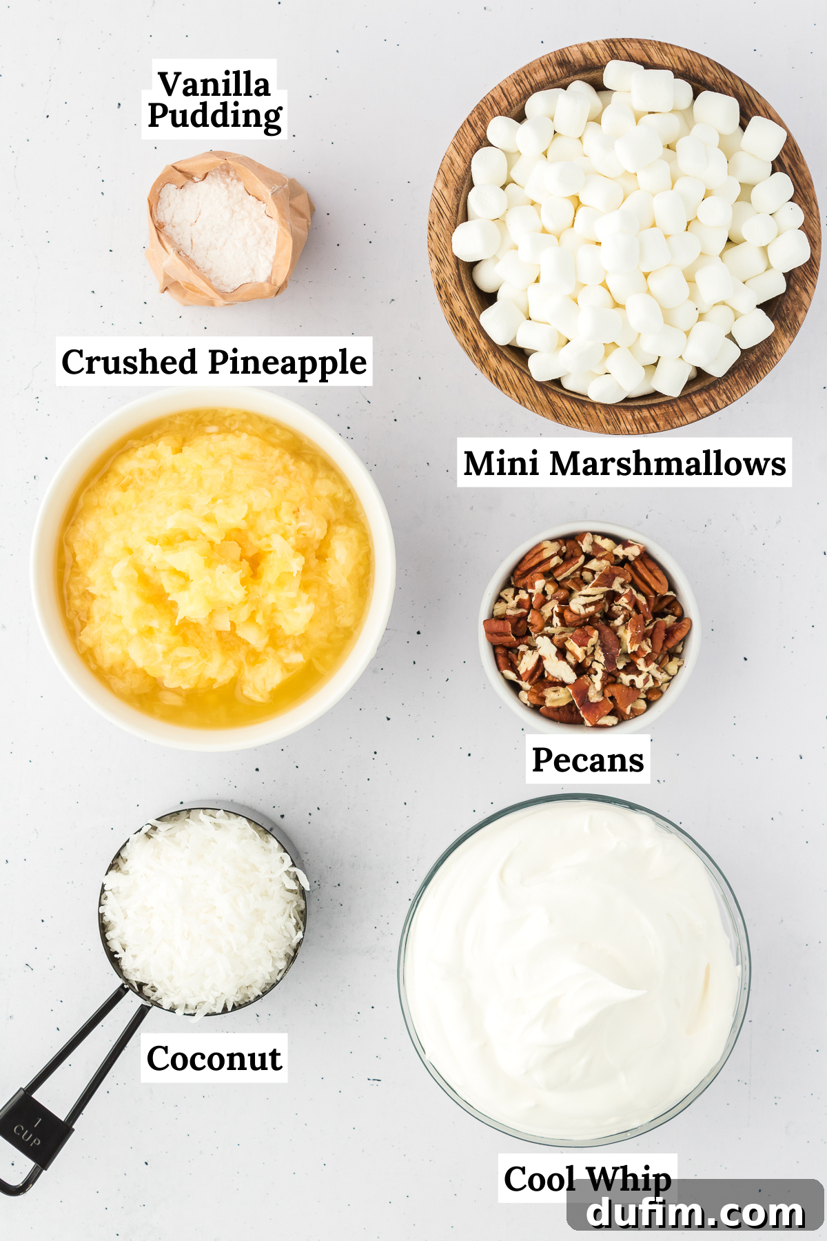 A vibrant display of all the essential ingredients for pineapple fluff: a box of instant vanilla pudding mix, a can of crushed pineapple, a tub of cool whip, a bag of mini marshmallows, a bowl of sweetened shredded coconut, and a small pile of chopped nuts.