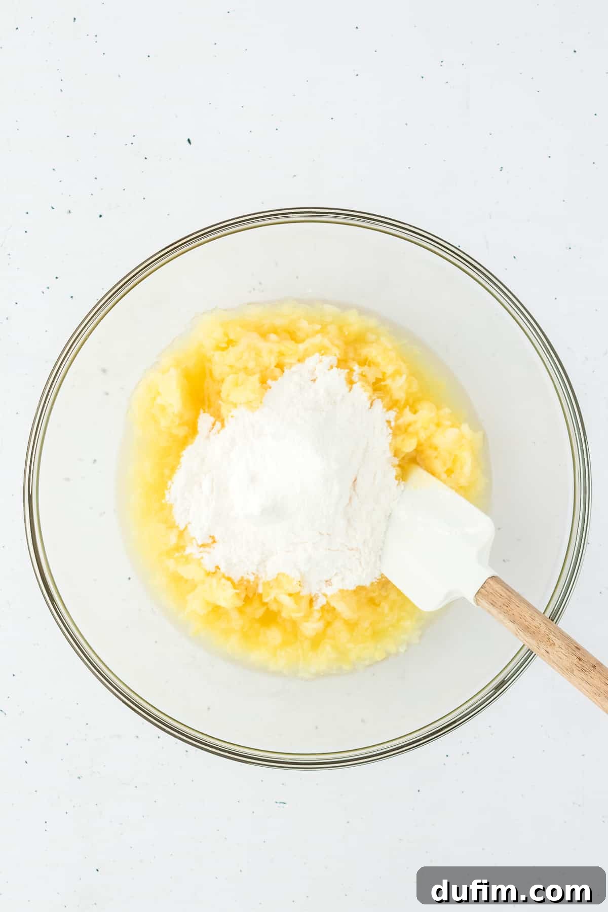 Crushed pineapple and vanilla pudding mix combined in a clear glass bowl, with a spatula resting inside, ready for mixing.