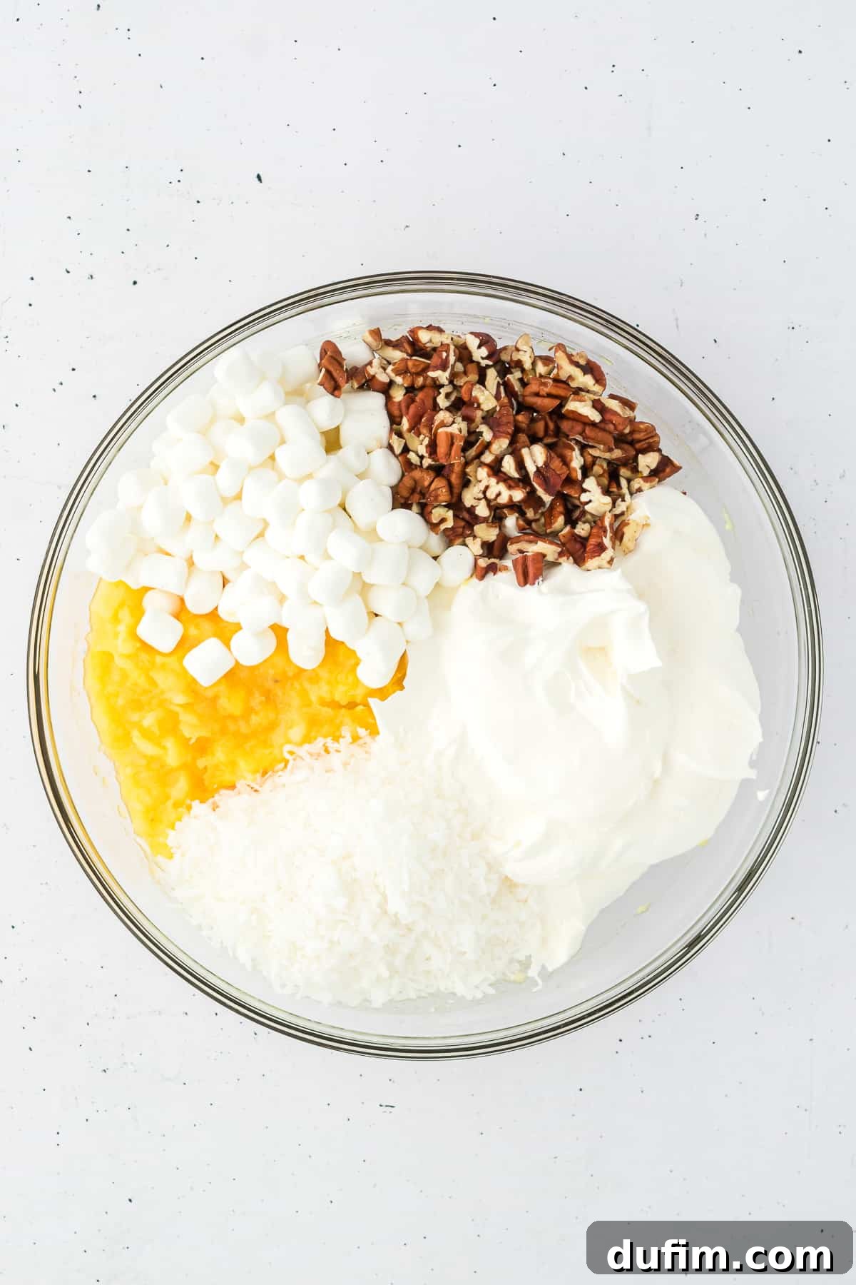 All the dry and wet ingredients for pineapple fluff – crushed pineapple, mini marshmallows, nuts, cool whip, and shredded coconut – gracefully mixed in a clear glass bowl.