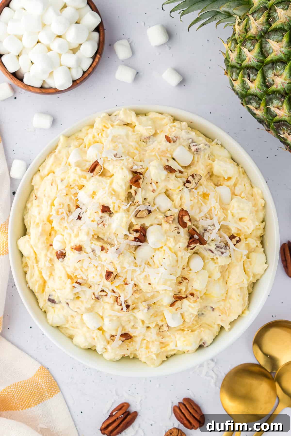 A pristine white bowl filled with creamy pineapple fluff, artfully arranged amidst a whole fresh pineapple, a bowl of mini marshmallows, kitchen towels, scattered nuts, and elegant gold spoons, highlighting its serving appeal.