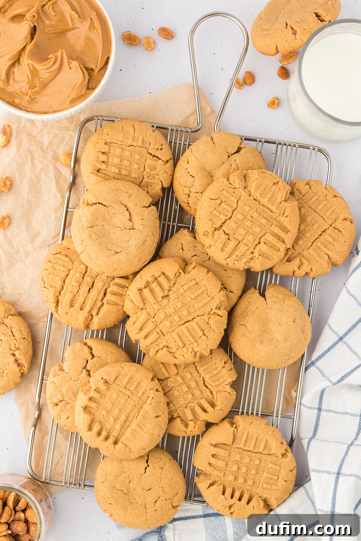 gluten free peanut butter cookies on a wire cooling rack surrounded by a white and blue striped kitchen towel, a bowl of peanuts, a bowl of peanut butter and a glass of milk