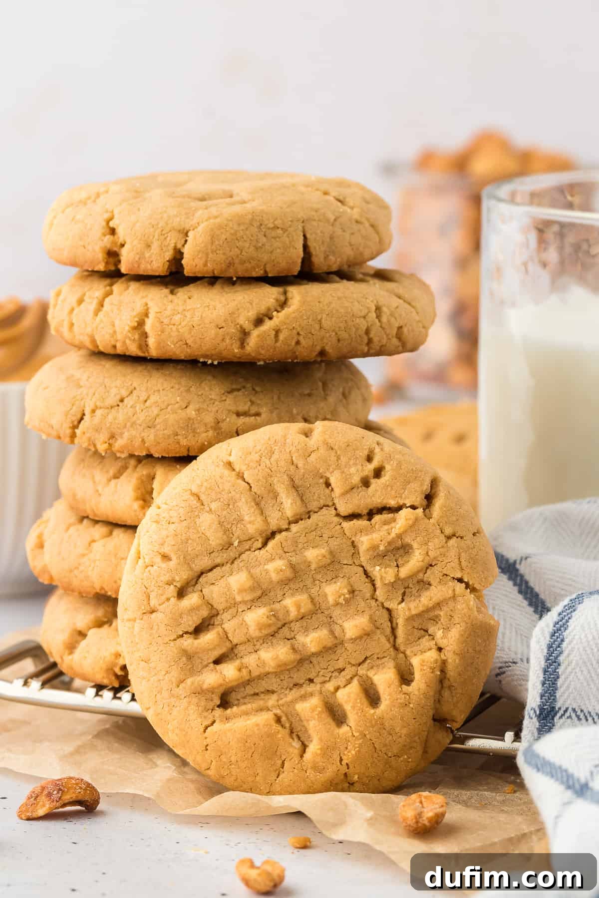 a stack of gluten free peanut butter cookies on a wire cooling rack with one cooking leaning vertically on the side of the stack, a white and blue kitchen towel, peanuts, and a glass of milk around them