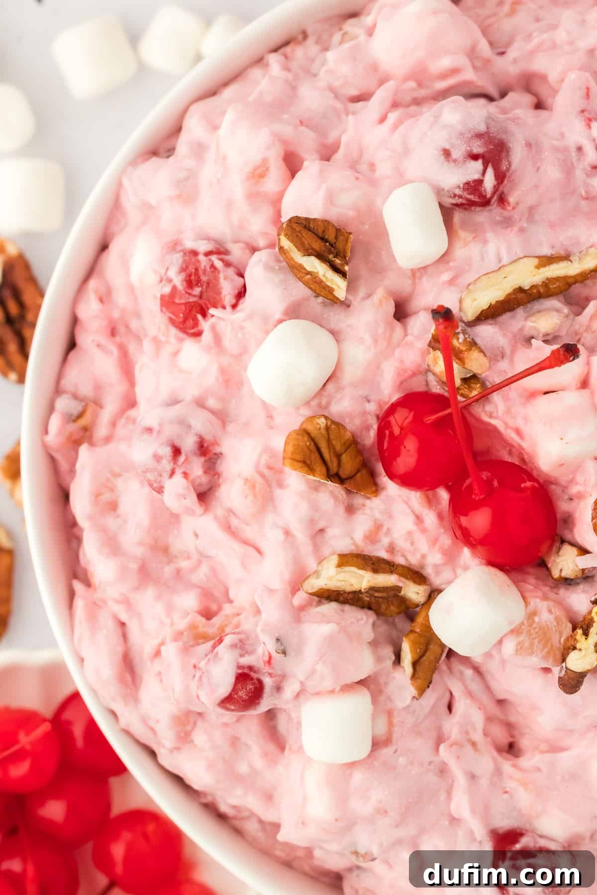 Close-up of a bowl of cherry fluff topped with maraschino cherries, mini marshmallows, and chopped pecans, surrounded by more cherries, nuts, and marshmallows