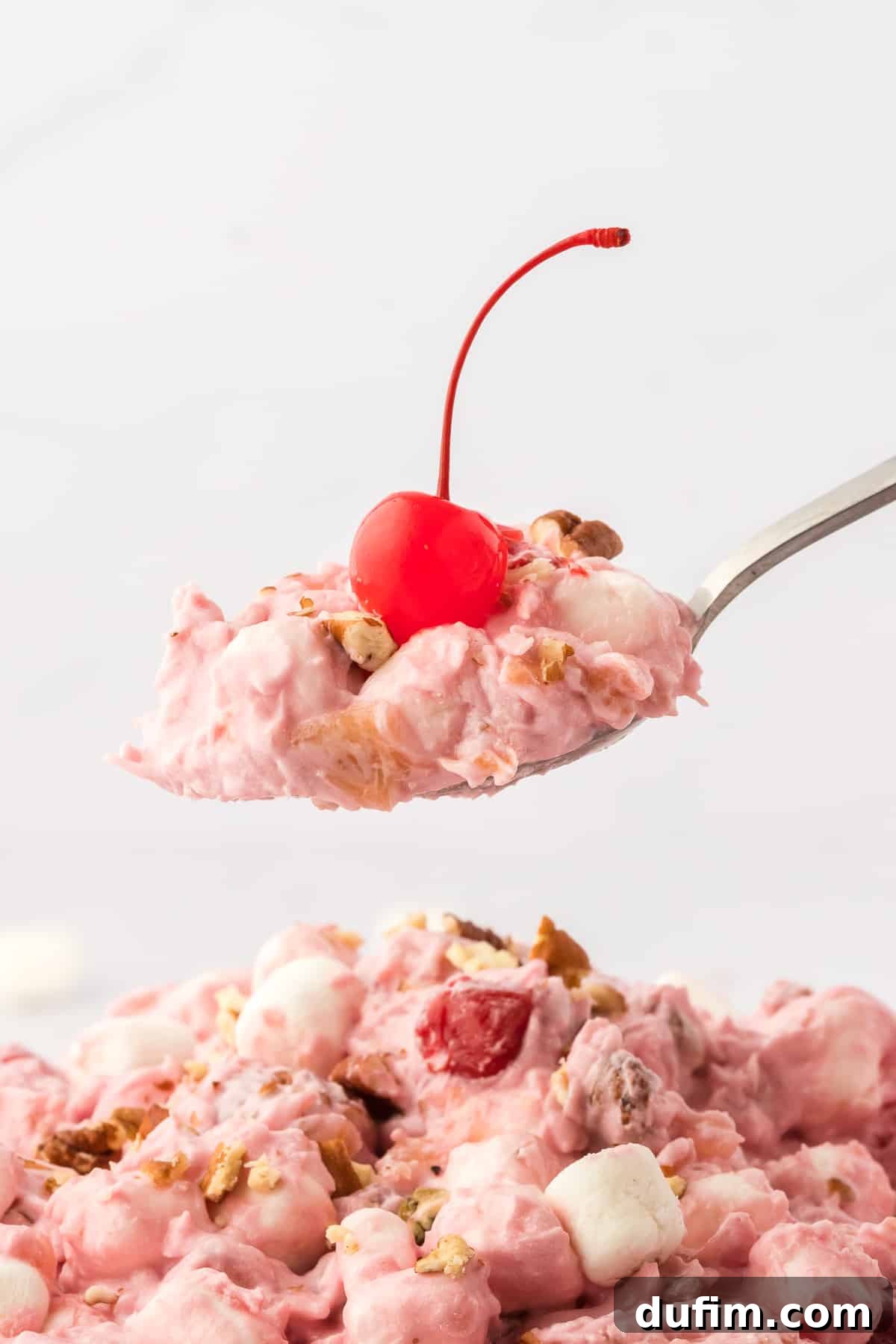 A spoon full of cherry fluff topped with a maraschino cherry held above more cherry fluff salad