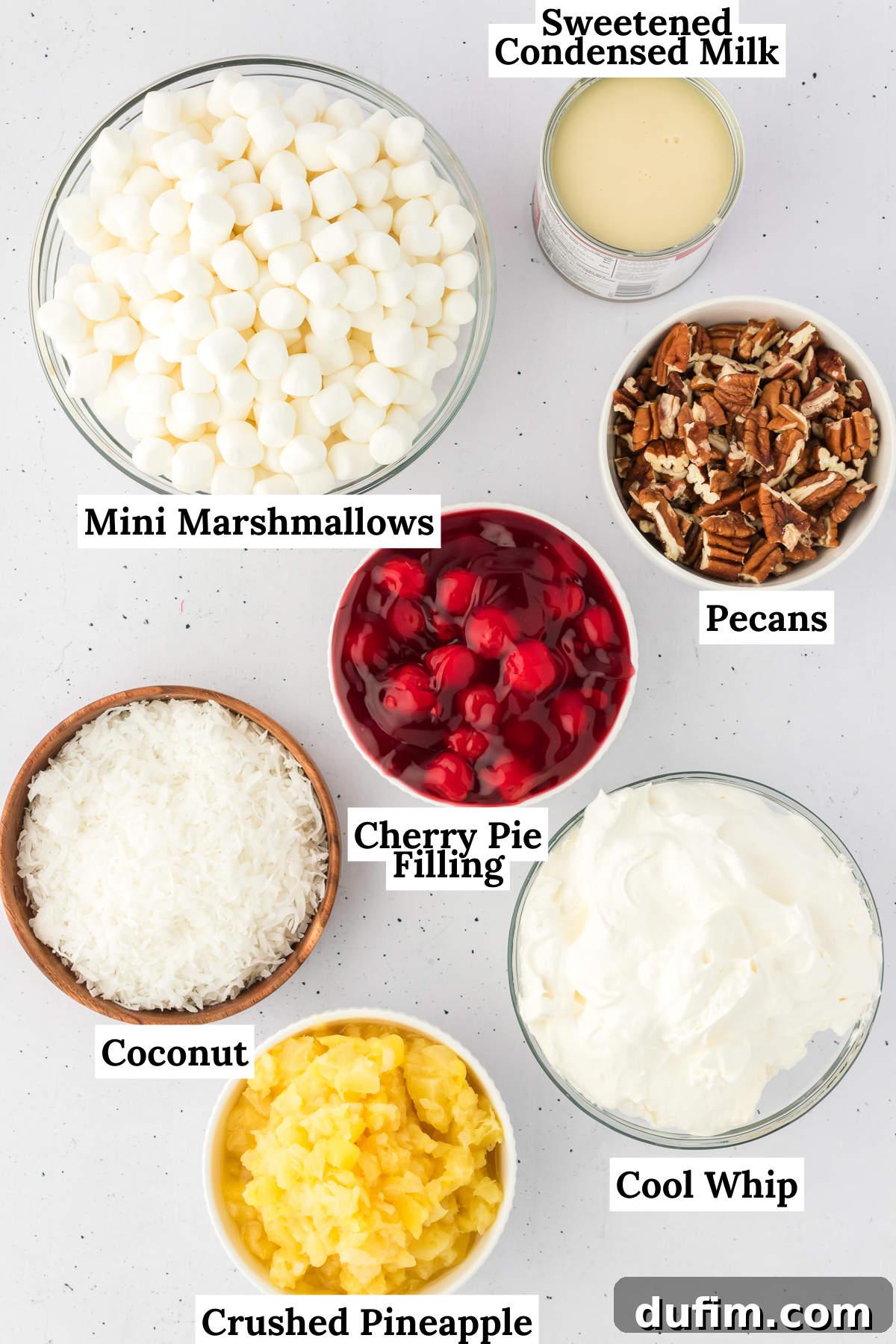 Ingredients for cherry fluff arranged on a flat surface including cherry pie filling, crushed pineapple, sweetened condensed milk, cool whip, sweetened shredded coconut, chopped pecans and mini marshmallows