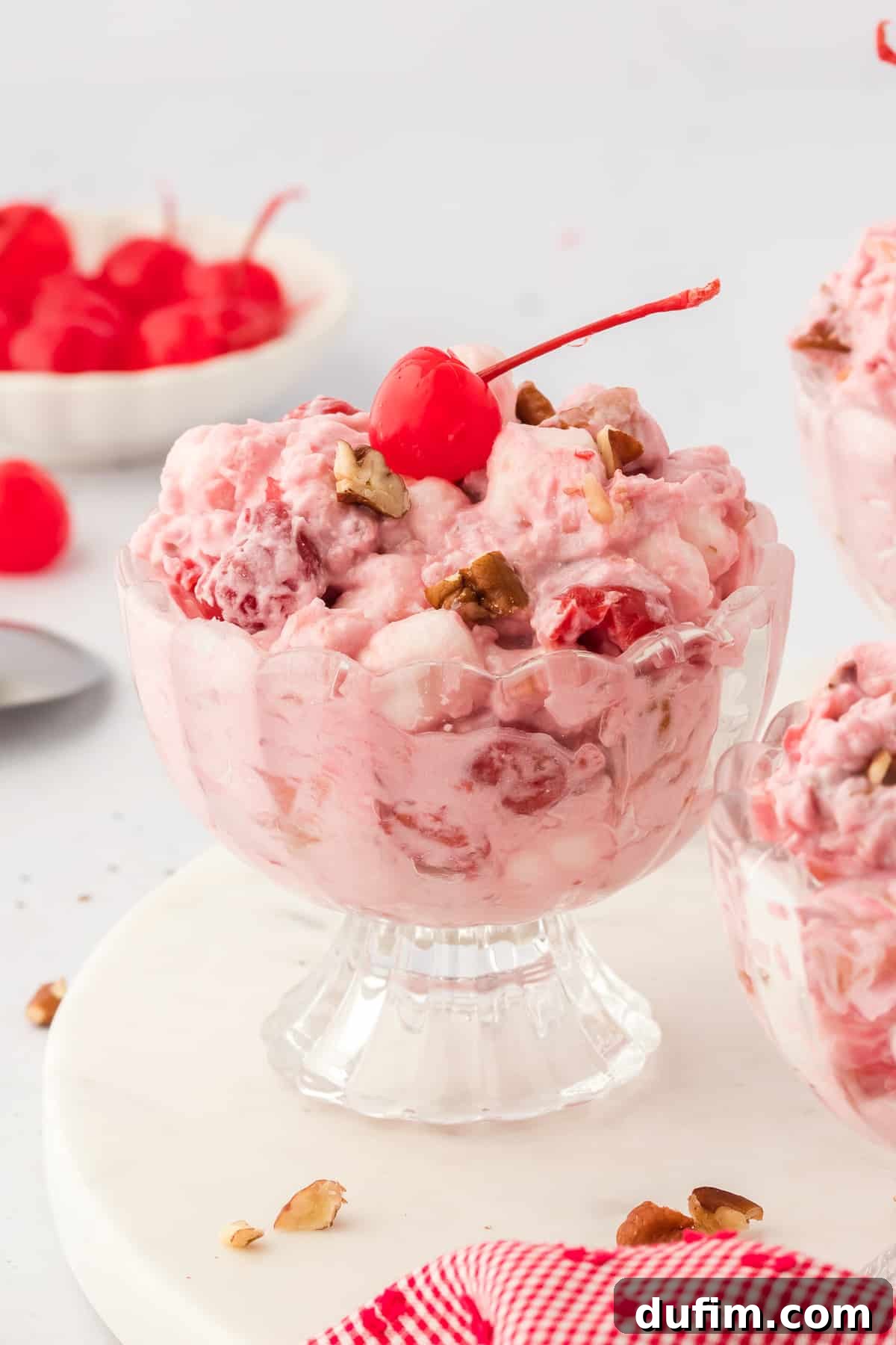 A glass bowl of cherry fluff topped with a maraschino cherry and surrounded by a bowl of cherries, more bowls of cherry fluff, and sprinkled nuts and a red and white towel around them