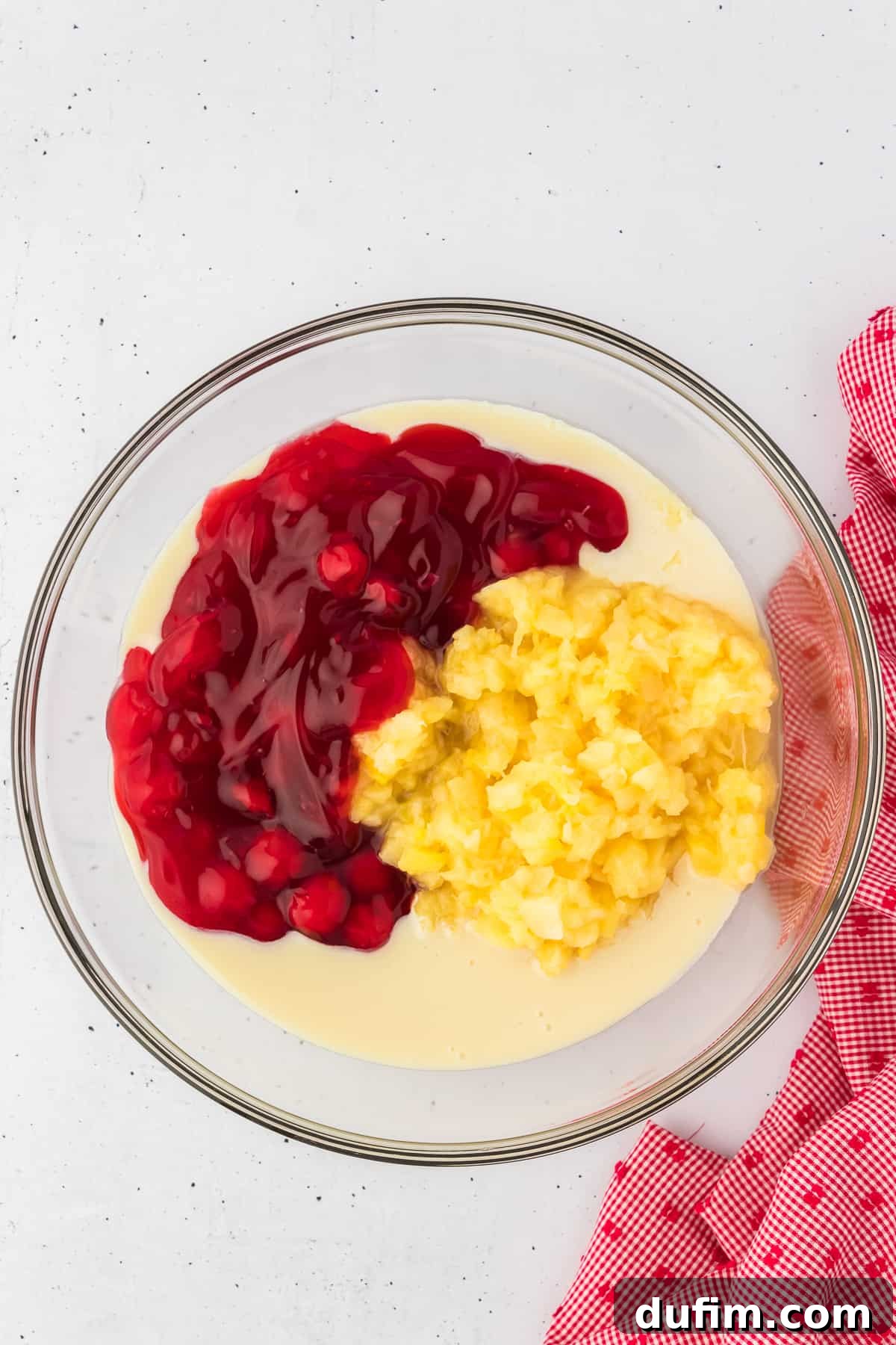 Condensed milk, cherry pie filling, and crushed pineapple in a clear glass bowl with a red and white kitchen towel to the right of it.