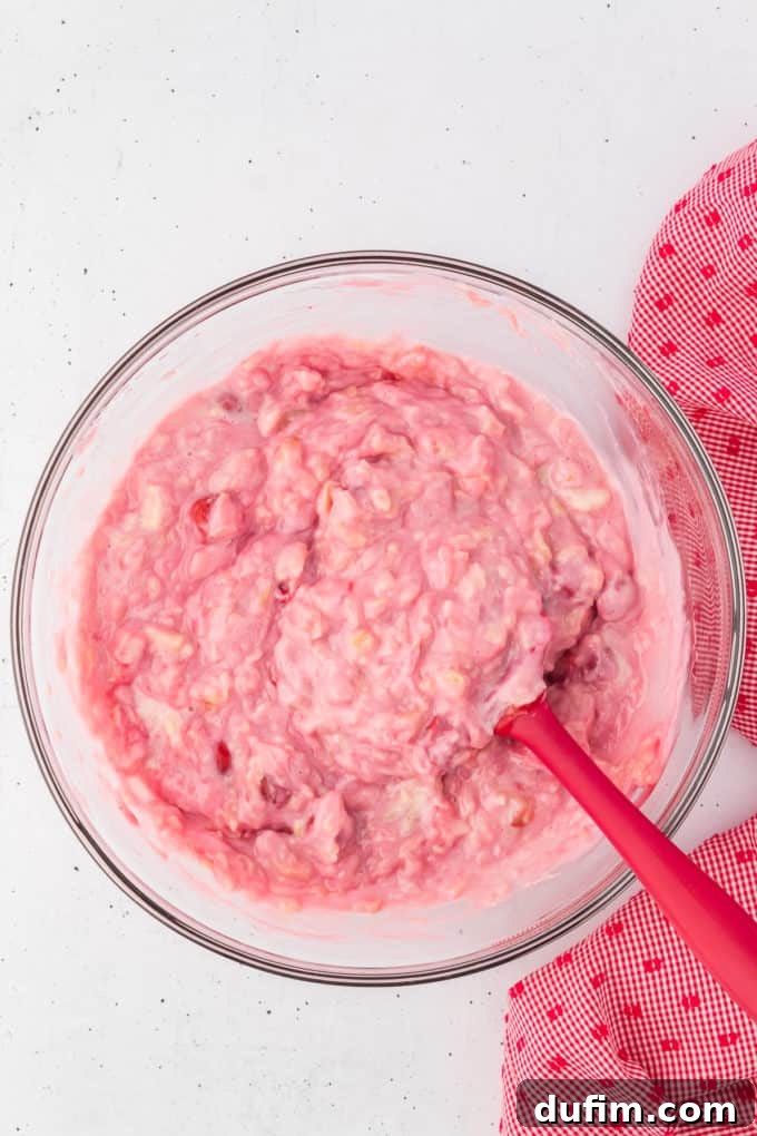 Ingredients for cherry fluff being mixed with a red spatula in a glass bowl with a red and white kitchen towel to the right of it.