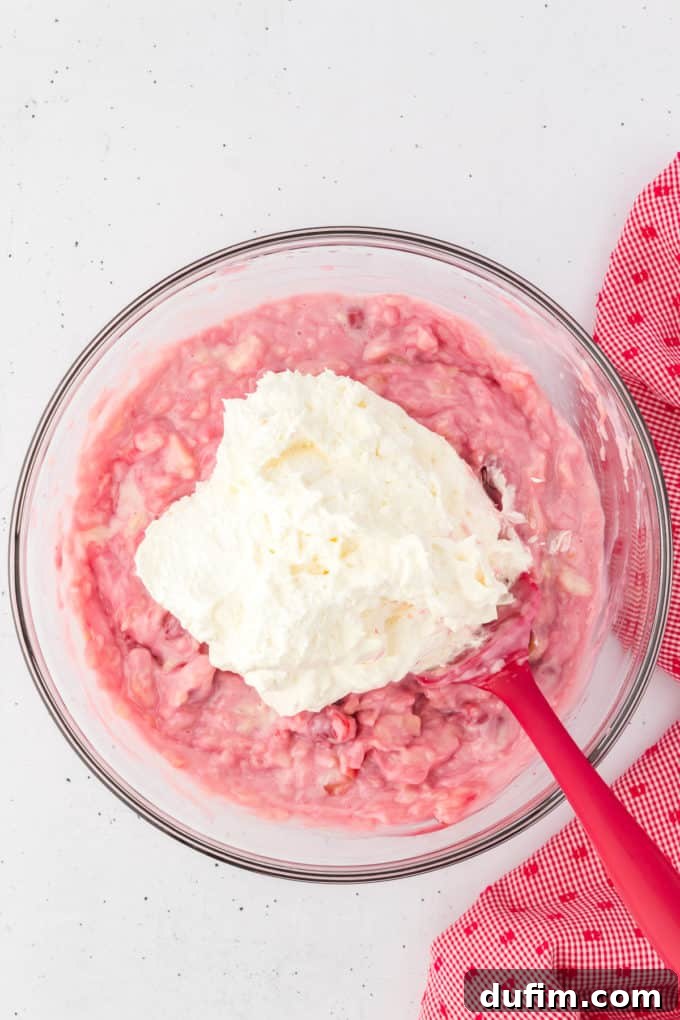 Cherry fluff ingredients mixed together with Cool Whip on top being mixed with a red spatula in a glass bowl with a red and white kitchen towel to the right of it.