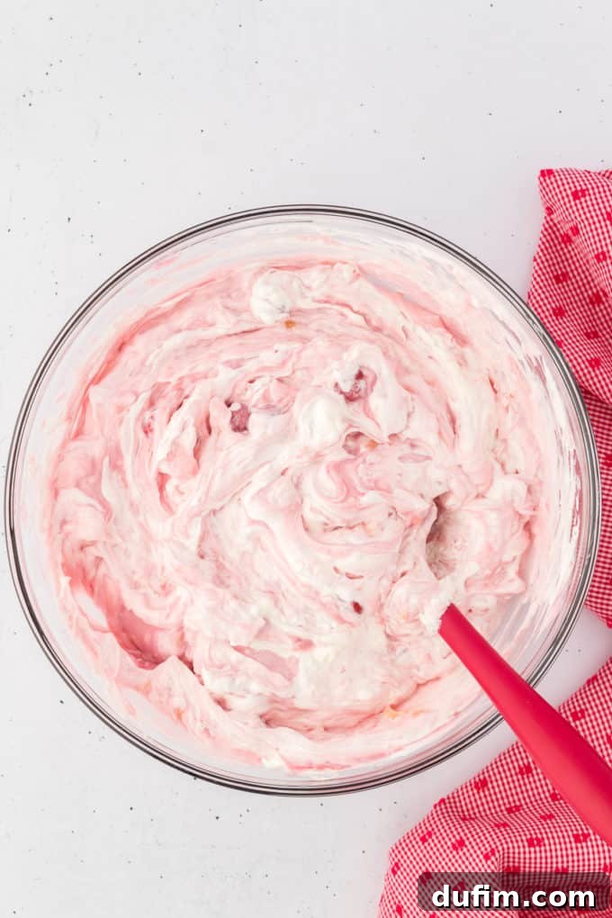 Cherry fluff ingredients mixed together with a red spatula in a glass bowl with a red and white kitchen towel to the right of it.