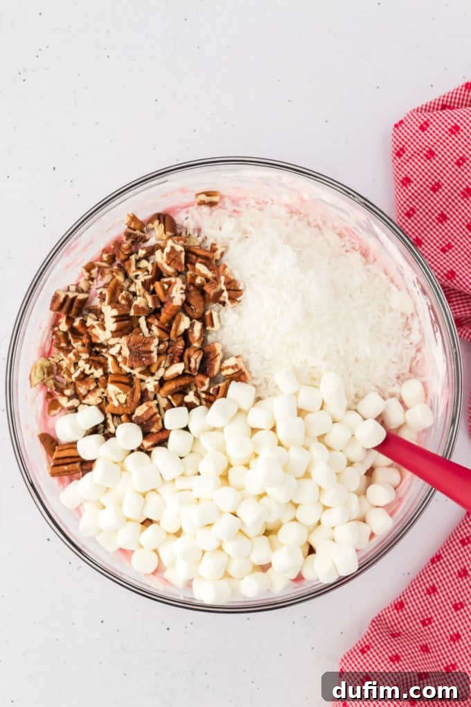 Cherry fluff ingredients in a glass bowl with chopped pecans, coconut, and marshmallows on top about to be mixed in with a red spatula, with a red and white kitchen towel to the right of it.