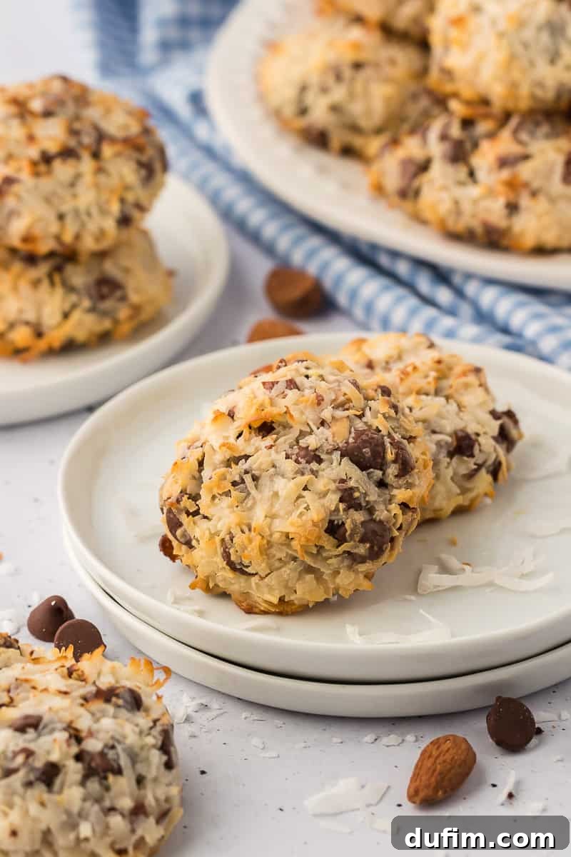almond joy cookies on a white plate with coconut flakes and chocolate chips sprinkled around, a blue and white checkered kitchen towel and more plates with almond joy cookies on them in the background