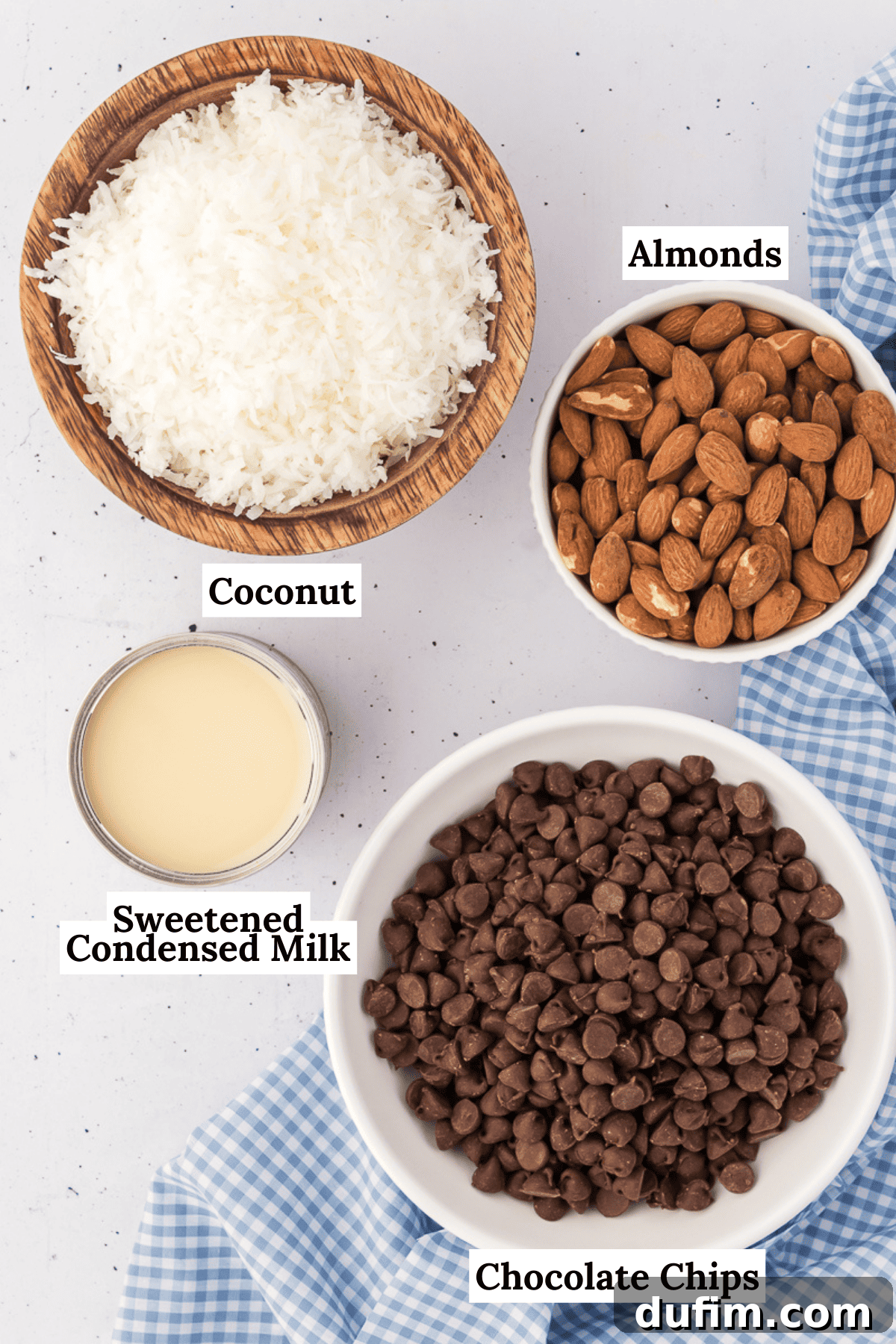 the ingredients for almond joy cookies including almonds, sweetened coconut flakes, semi-sweet chocolate chips, salt and sweetended condensed milk with a blue and white checkered kitchen towel