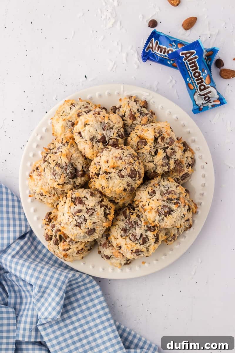A pile of almond joy cookies on a white plate with two almond joy candy bars beside it and coconut flakes, almonds and chocolate chips sprinkled around