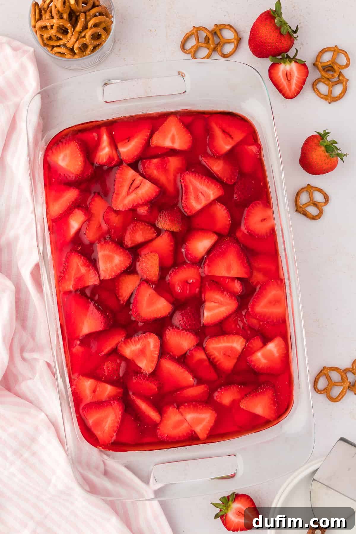 A clear glass baking dish of strawberry pretzel salad surrounded by fresh strawberries, pretzels, and a pink and white striped kitchen towel