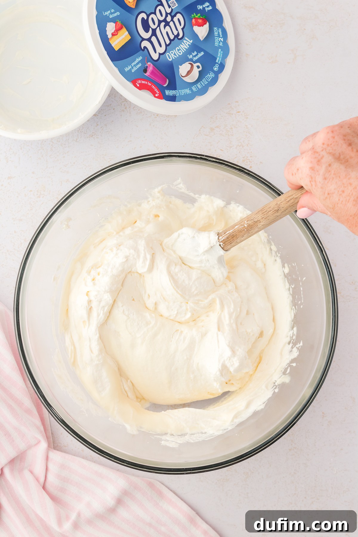Cream cheese mixture in a glass bowl being mixed with a spatula, beside a pink and white striped kitchen towel and an open container of cool whip