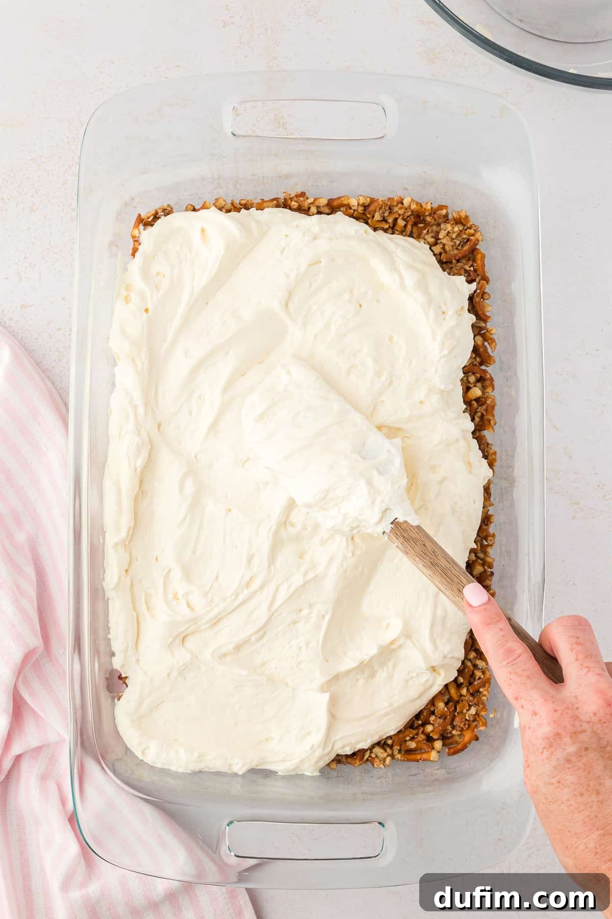 The cream cheese layer of strawberry pretzel salad being spread over top of the pretzel crust layer in a large glass baking dish, beside a pink and white kitchen towel