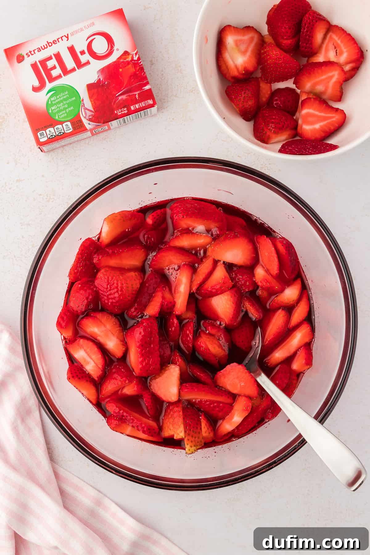 A glass bowl with the strawberry jello mixture in it for strawberry pretzel salad with a spoon in it, beside a small white bowl of fresh strawberries, a pink and white striped kitchen towel and a box of strawberry jello