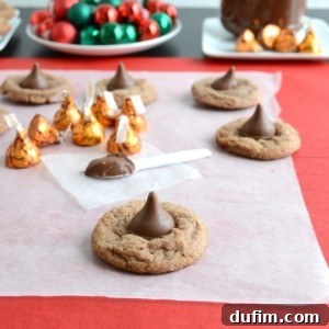 Chocolate cookies topped with Hershey’s Kisses are arranged on parchment paper, with unwrapped Kisses and a jar of chocolate spread in the background, alongside a plate of red and green holiday ornaments.