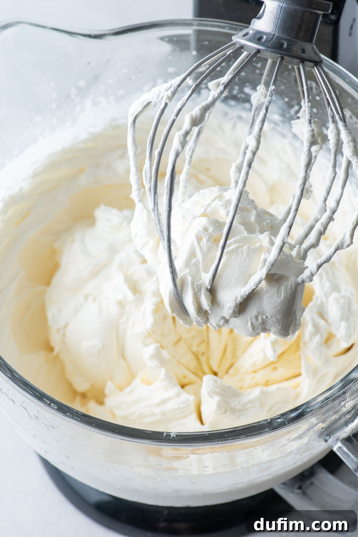 An electric mixer with the beater lifted, showcasing a large amount of perfectly mixed, stabilized whipped cream clinging to it.