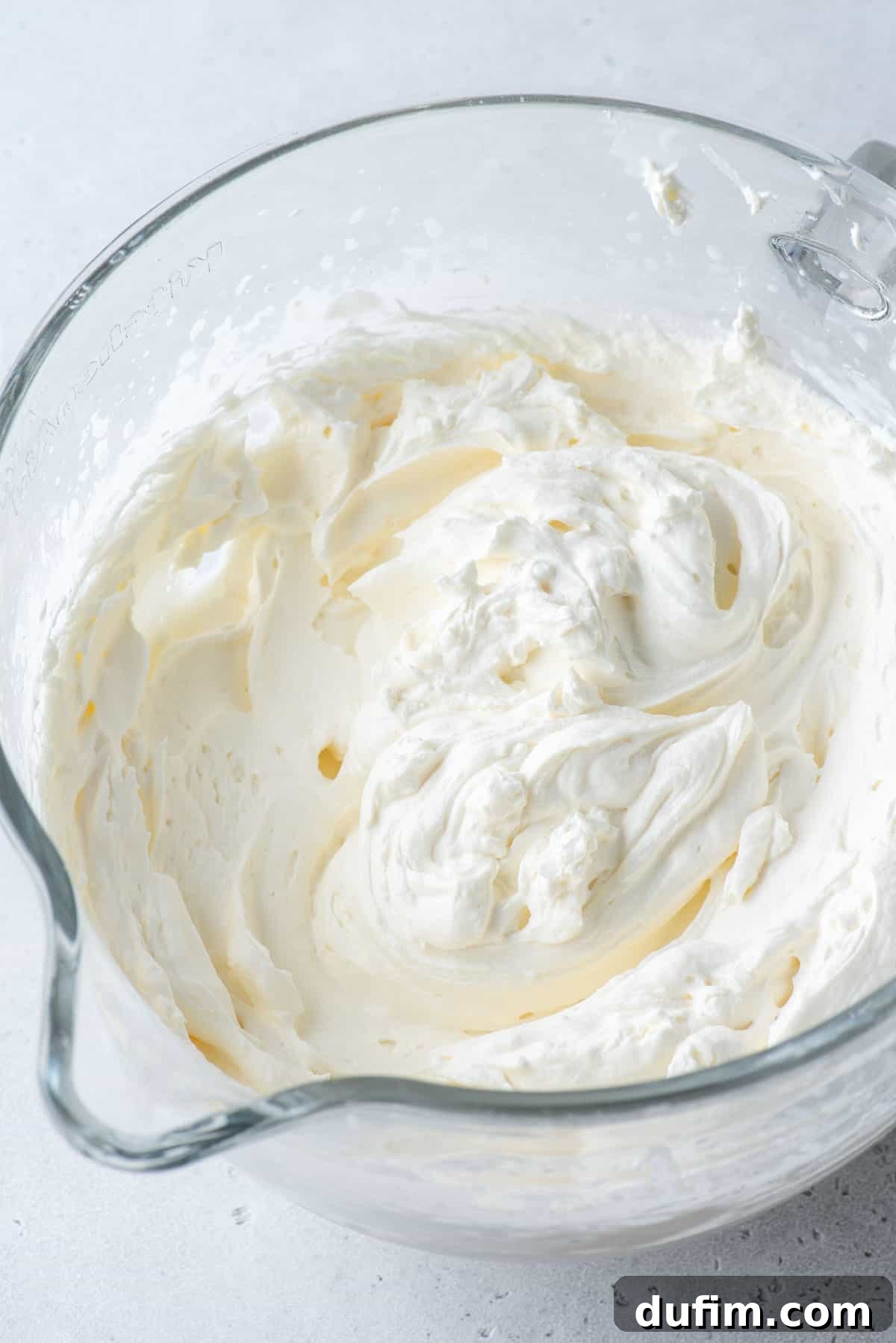 Stabilized whipped cream filling a clear glass mixing bowl, showing its ample volume and smooth texture.