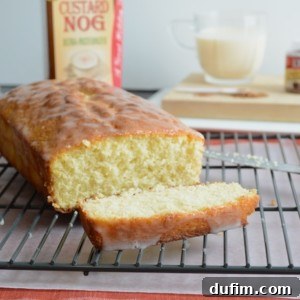 A loaf of glazed bread with one slice cut sits on a cooling rack. In the background, there is a carton of custard nog and a glass cup filled with a light-colored beverage.