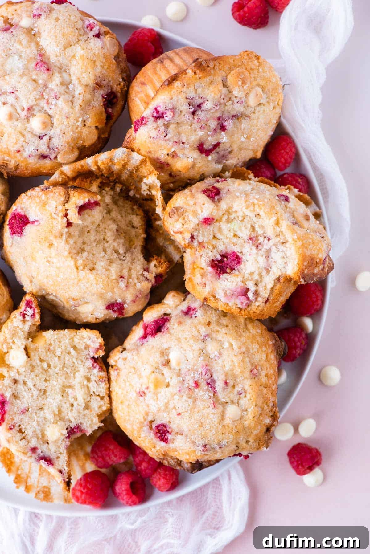 Decadent Raspberry White Chocolate Muffins 2 A beautifully arranged white plate brimming with fluffy raspberry white chocolate chip muffins, resting on a soft pink surface. The scene is artfully scattered with fresh, vibrant raspberries and delicate white chocolate chips, creating an enticing visual display.