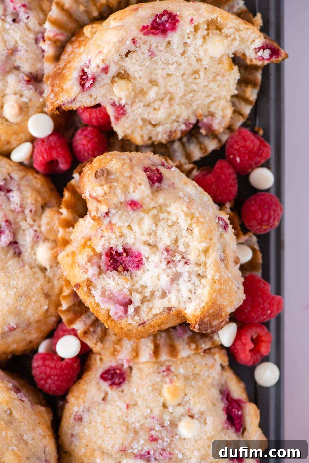 Decadent Raspberry White Chocolate Muffins 4 A vibrant display of a raspberry white chocolate muffin, sliced in half to reveal its tender, fruit-studded interior, rests atop a pile of whole muffins. Fresh, glistening raspberries and creamy white chocolate chips are scattered around, inviting a closer look at these delectable treats.
