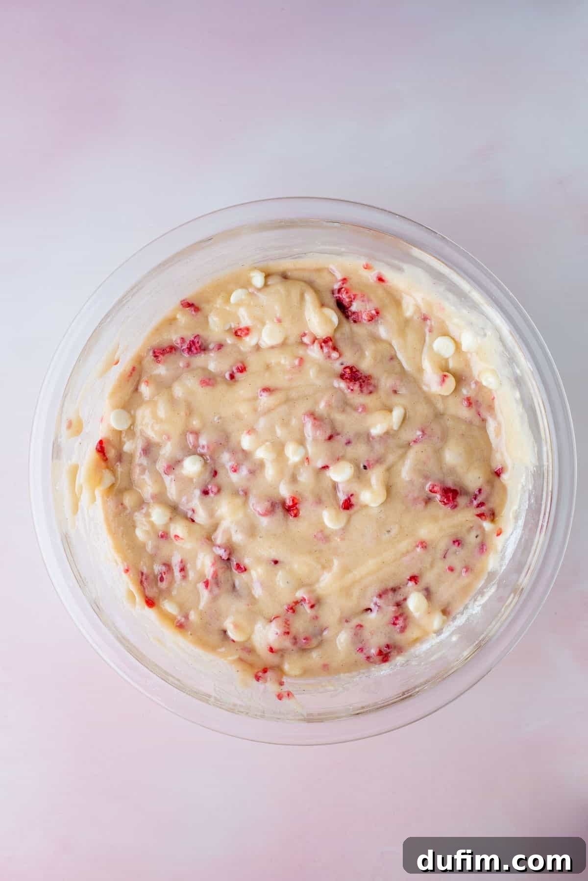 Decadent Raspberry White Chocolate Muffins 5 A pristine glass bowl holds a generous portion of luscious raspberry white chocolate chip muffin batter, poised on a charming pink surface, ready for baking.