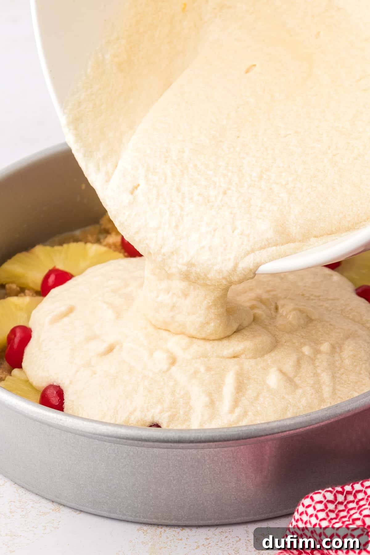 Thick pineapple upside-down cake batter is being carefully poured over the artfully arranged pineapple and cherry topping in a prepared cake pan.