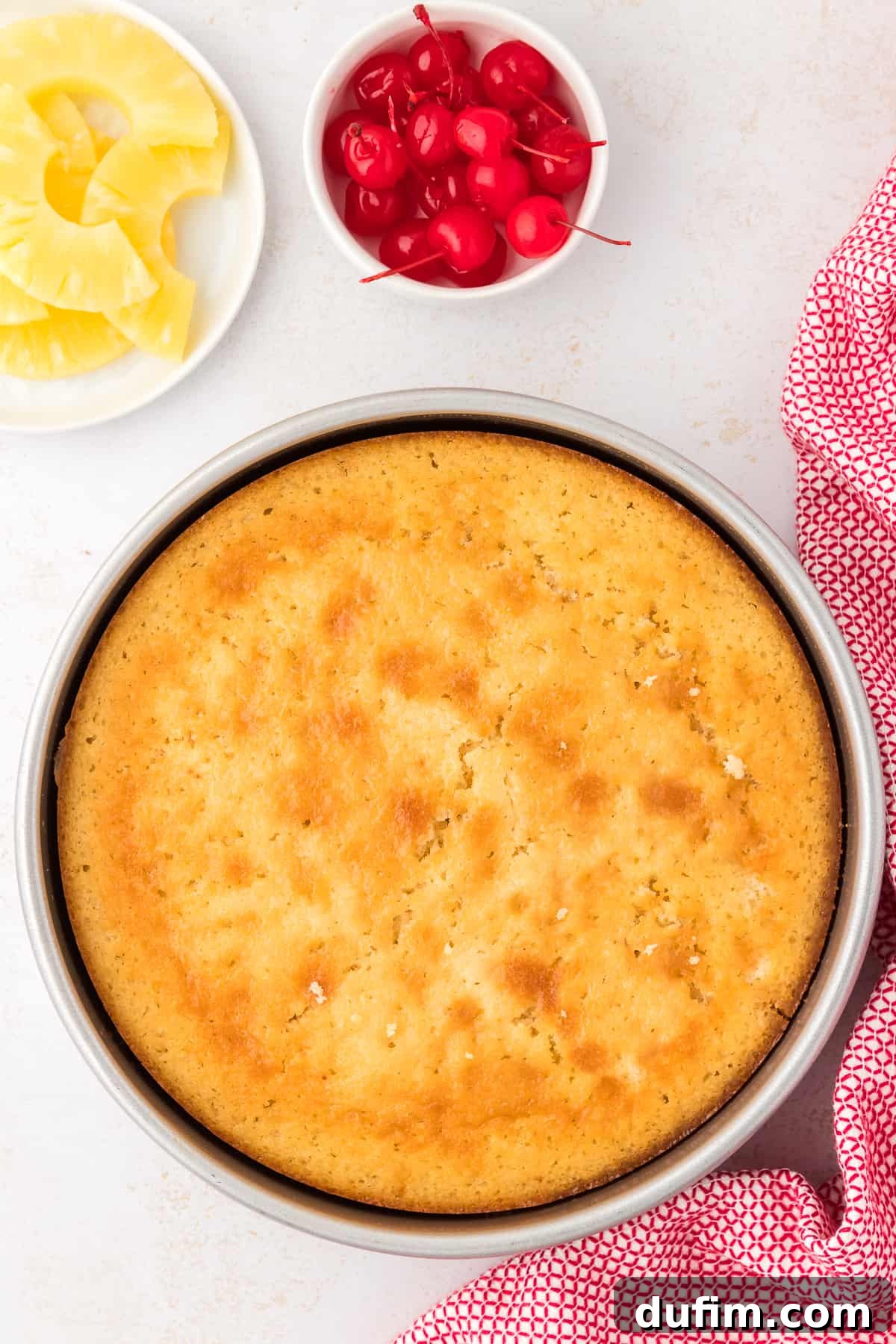 The pineapple upside-down cake, fresh out of the oven and beautifully golden, resting in its pan before being inverted, with garnishes like pineapple slices and cherries in the background.