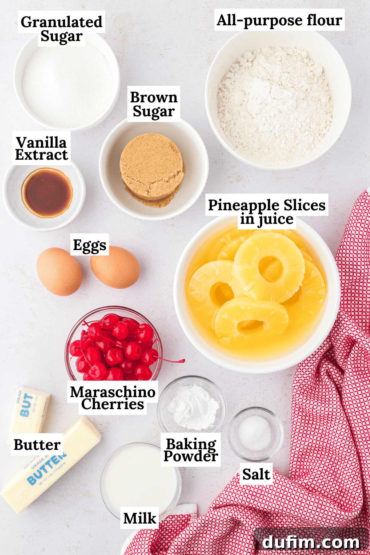 A vibrant flat lay of all the essential ingredients for pineapple upside down cake, including golden pineapple slices, bright red maraschino cherries, various sugars, and baking staples.