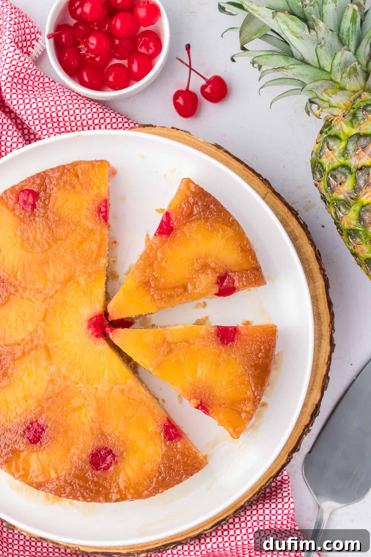 A beautifully sliced pineapple upside-down cake on a white plate, ready to be served, surrounded by decorative elements like a red and white kitchen towel and a serving spatula.
