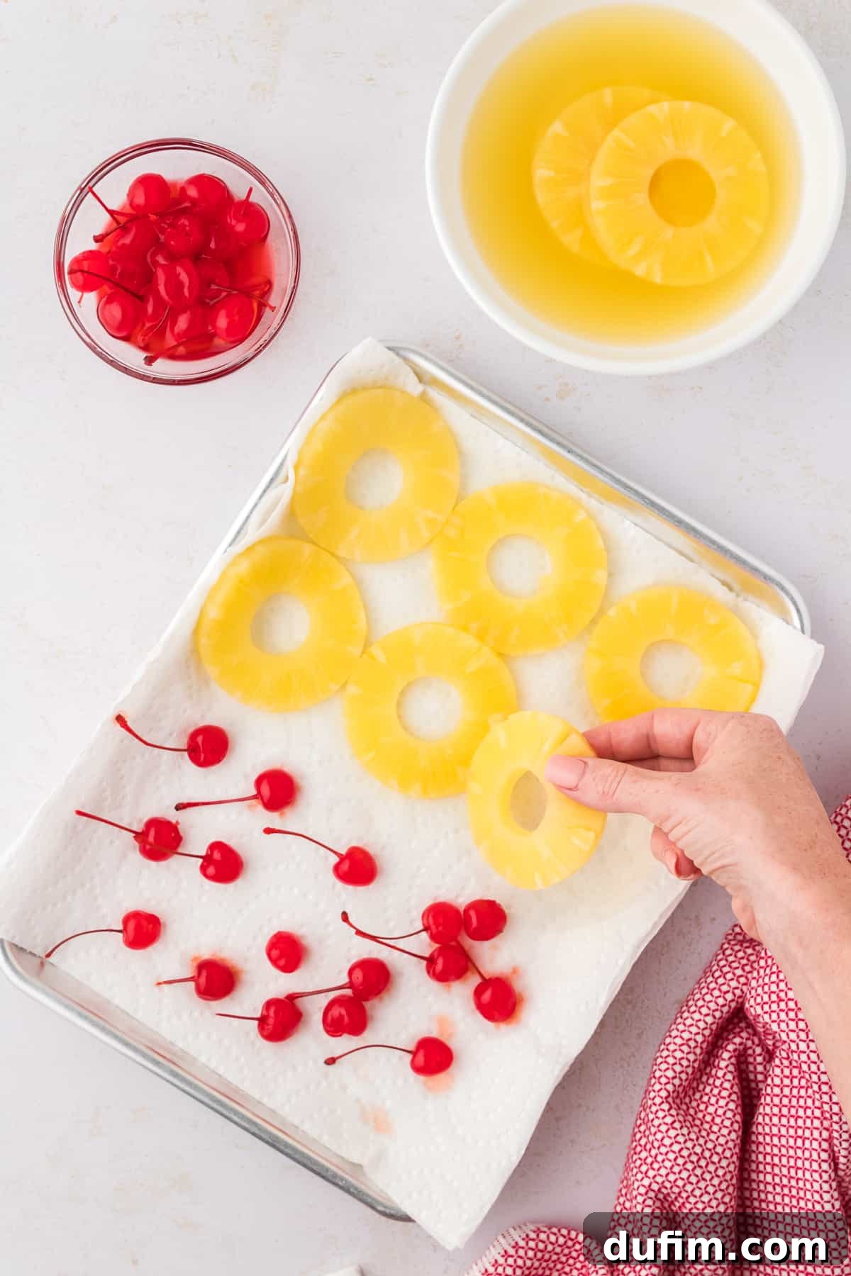 Pineapple slices and maraschino cherries are laid out on paper towels on a baking sheet to blot excess moisture, with bowls of fresh fruit nearby.