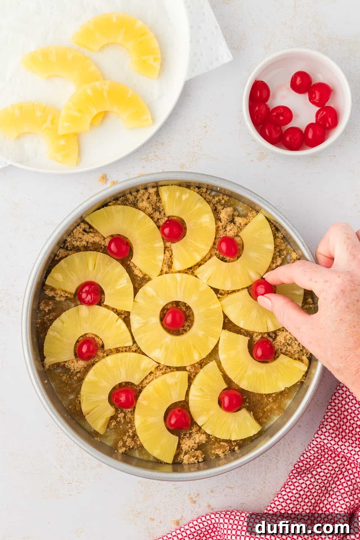 A hand carefully arranging vibrant pineapple slices and maraschino cherries in a decorative pattern over the brown sugar and melted butter base in a cake pan, creating the beautiful upside-down design.