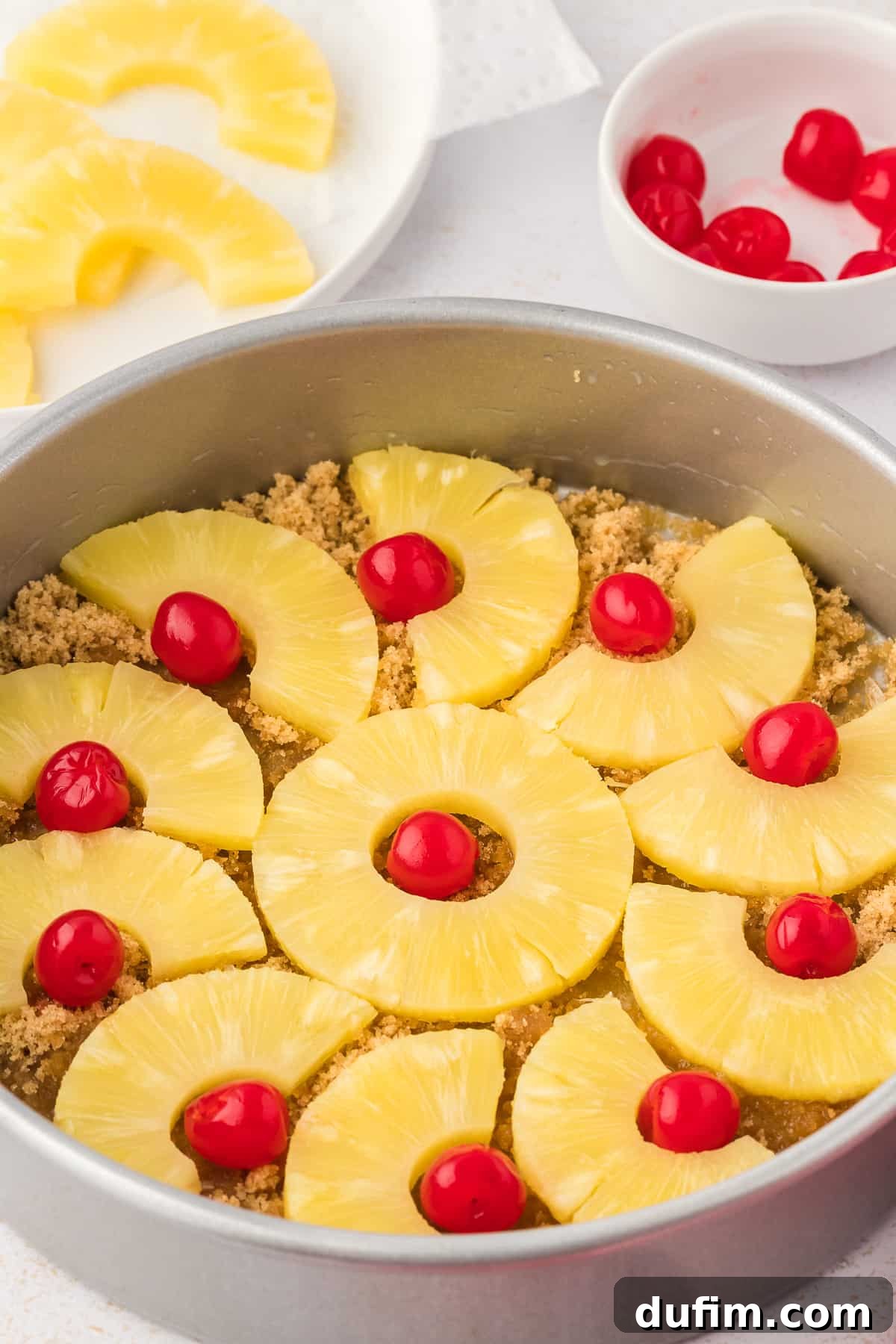 The fully arranged base of the pineapple upside-down cake in the pan, showcasing the intricate pattern of pineapple slices and maraschino cherries over the caramelized brown sugar and butter layer, ready for the batter.