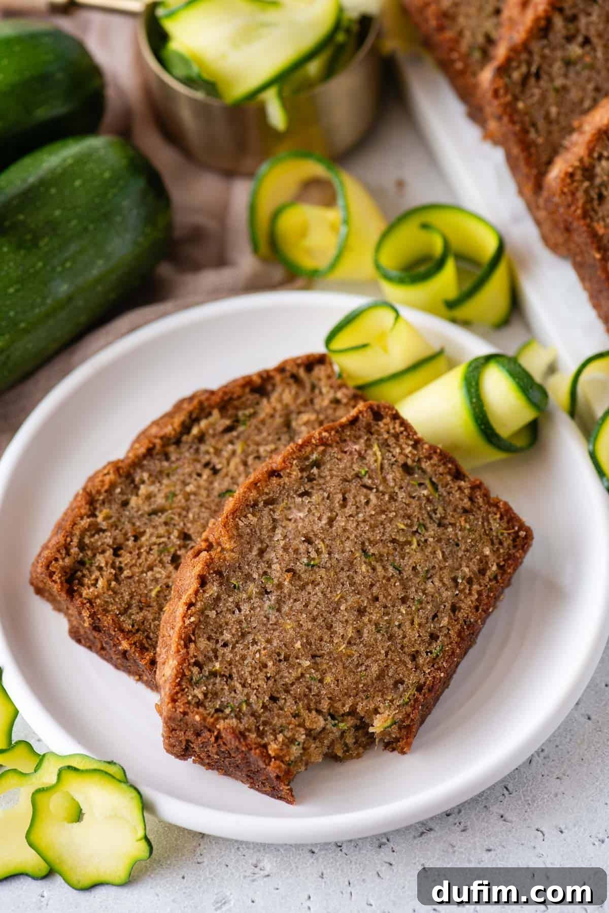 Irresistibly Moist Zucchini Bread 2 Two slices of perfectly moist zucchini bread on a white plate, surrounded by fresh sliced zucchini and whole zucchinis, highlighting its natural goodness.